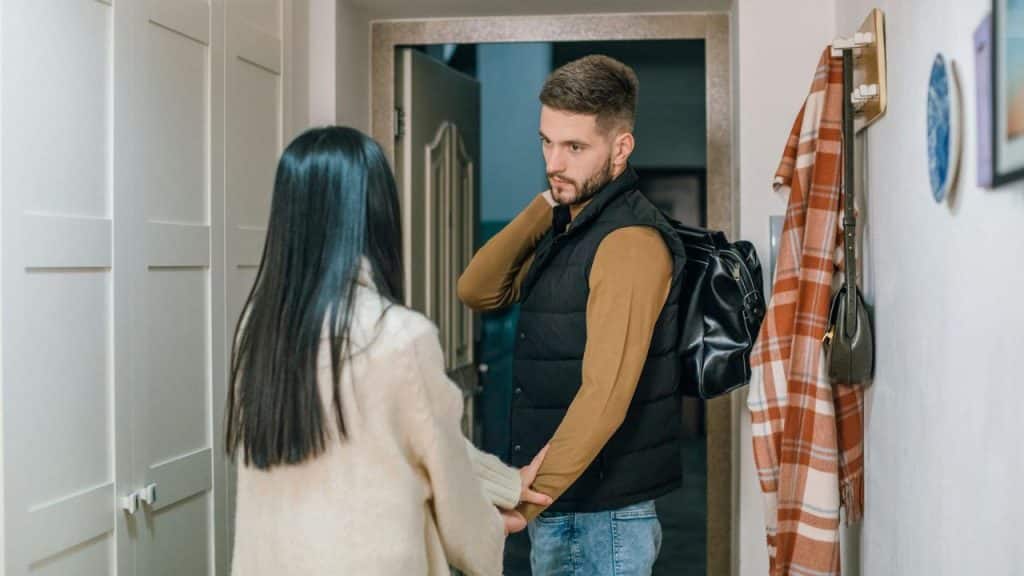 A man carrying a bag stands by a doorway while a woman holds his arm.