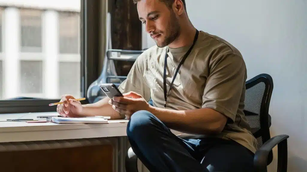 A man sitting at a desk using his phone while holding a pencil.