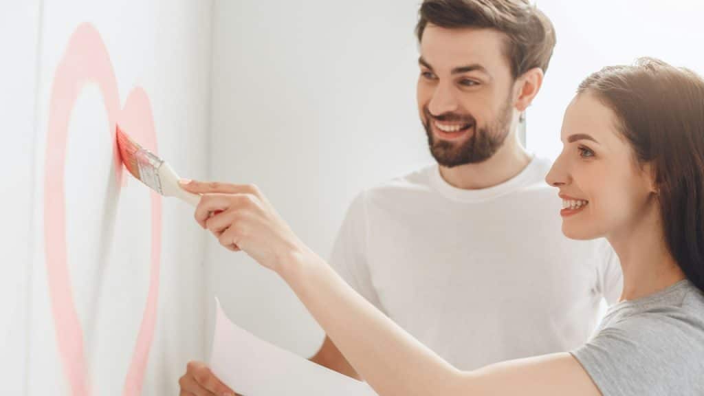 A woman is painting a heart on a wall while her husband, holding a piece of paper, is standing next to her.