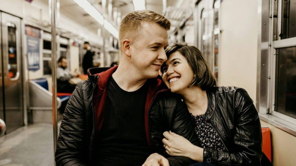 A man and woman sit closely together on a train, looking at one another.