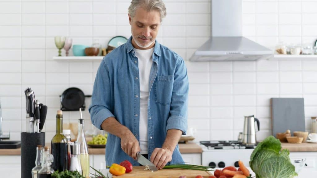 A man in a denim shirt chops green onions in a bright, modern white kitchen.