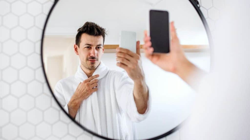 A man in a white robe takes a selfie in a circular mirror with honeycomb tiles.