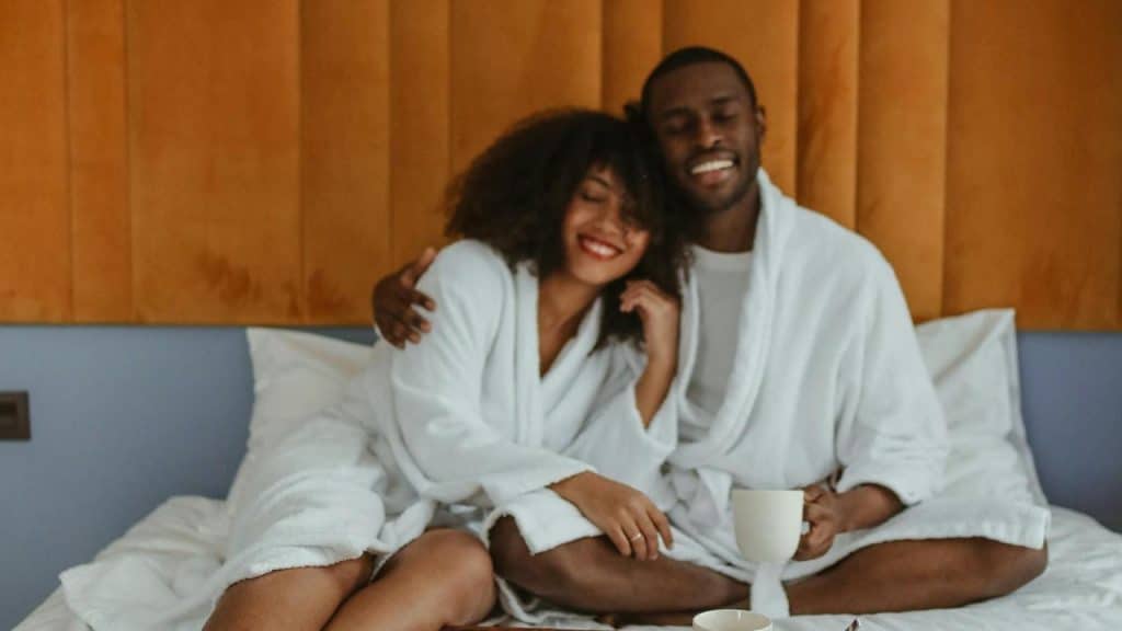A couple in white robes sitting on a bed and smiling warmly together.