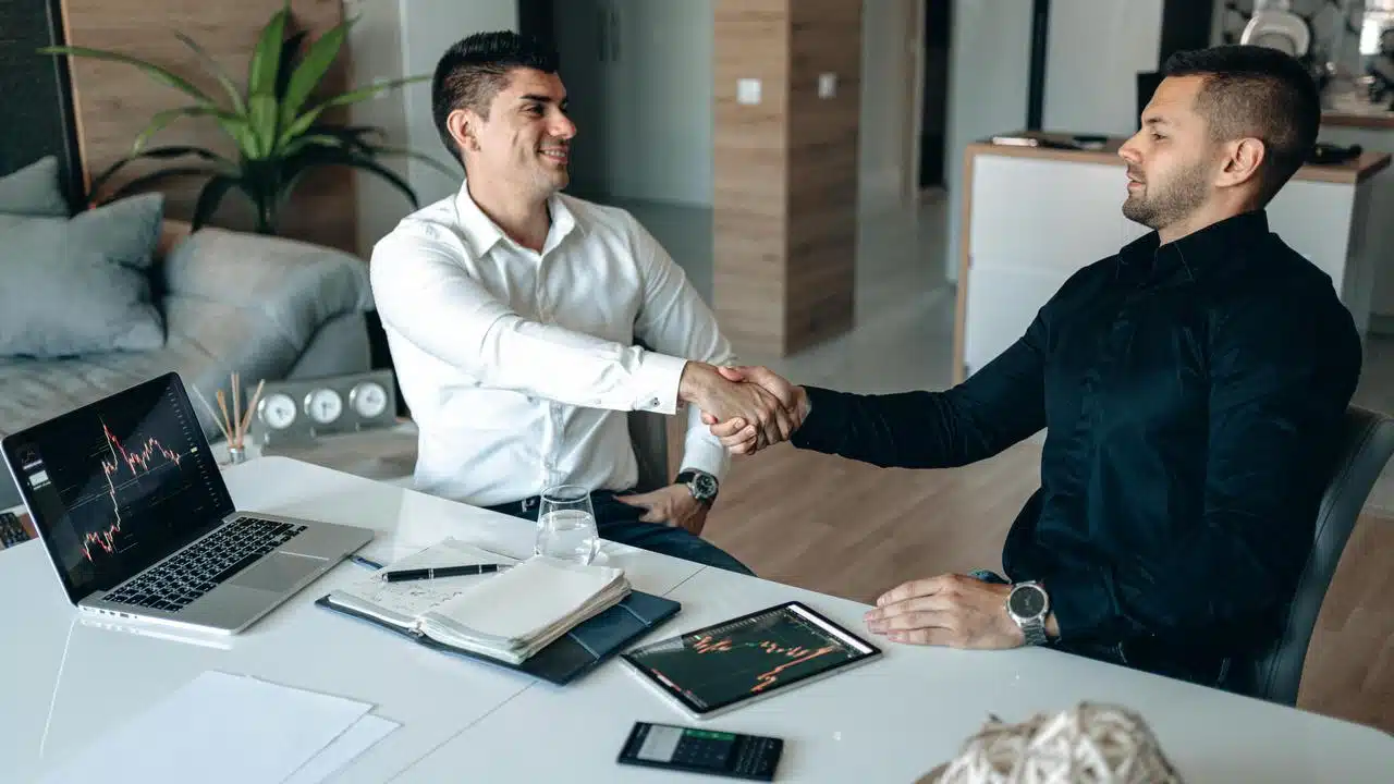 A handshake between two businessmen sitting at a desk in an office.