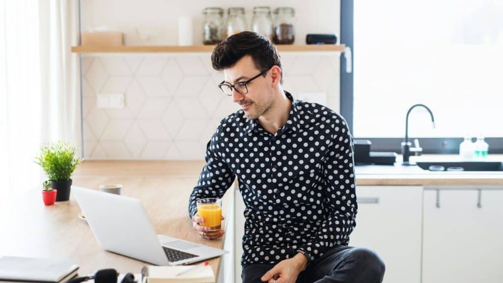 A man wearing a polka-dot shirt sits in a kitchen while looking at a laptop.