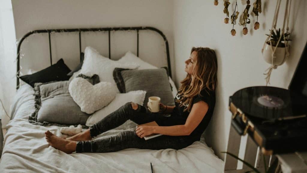 A woman in a black t-shirt and jeans sits on a bed, leaning against the wall with a mug and notebook.