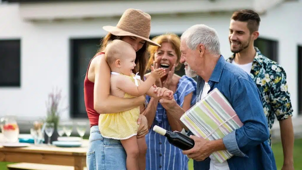 A family gathers outdoors while an older man holds a gift and reaches for a baby.