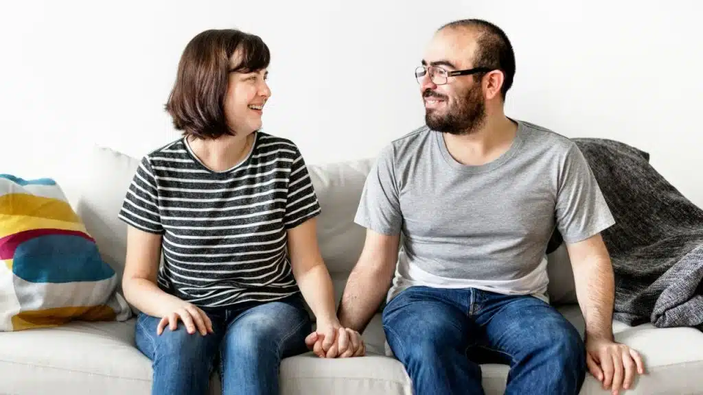 A smiling man and woman sit on a gray sofa holding hands and looking at each other.