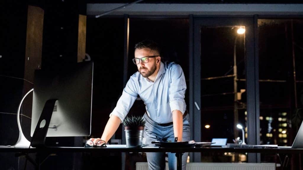A man with glasses standing at a desk in a dark office looking at a computer.