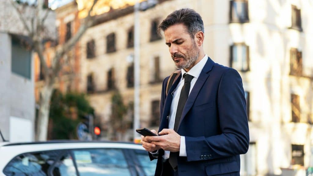 A man in a suit stands on a city street looking down at his phone.