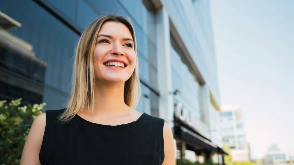 A blonde woman in a black top smiles while looking toward the sky outside buildings.