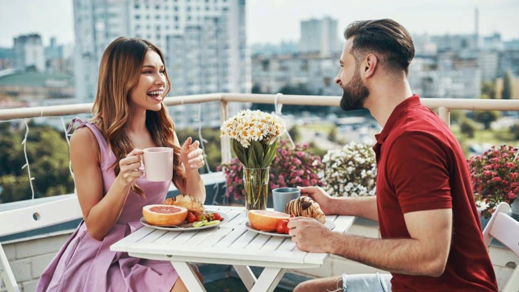 A man and woman sit at a white table on a balcony having breakfast together.
