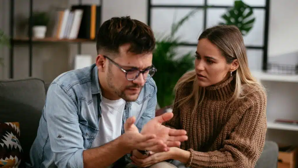 A man with glasses gestures with his hands while a woman in a sweater looks at him.