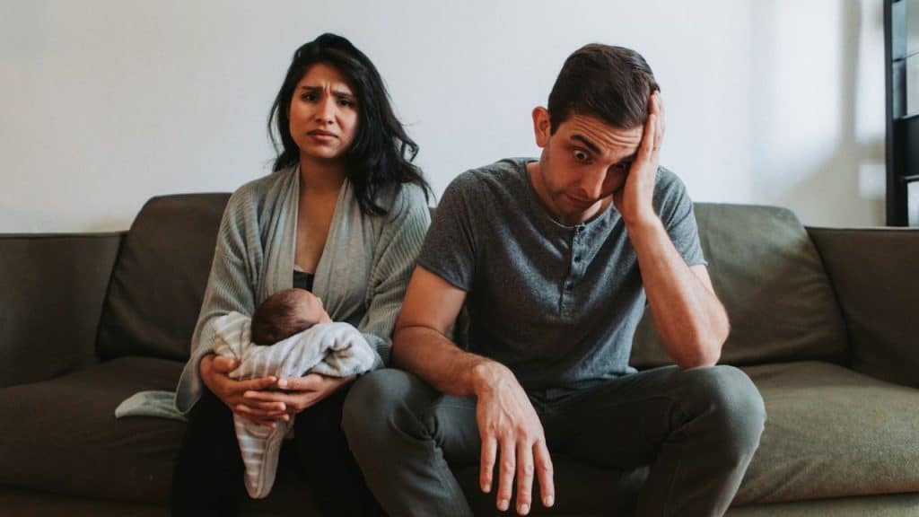 A woman holds a baby while a man beside her sits with his hand on his head.