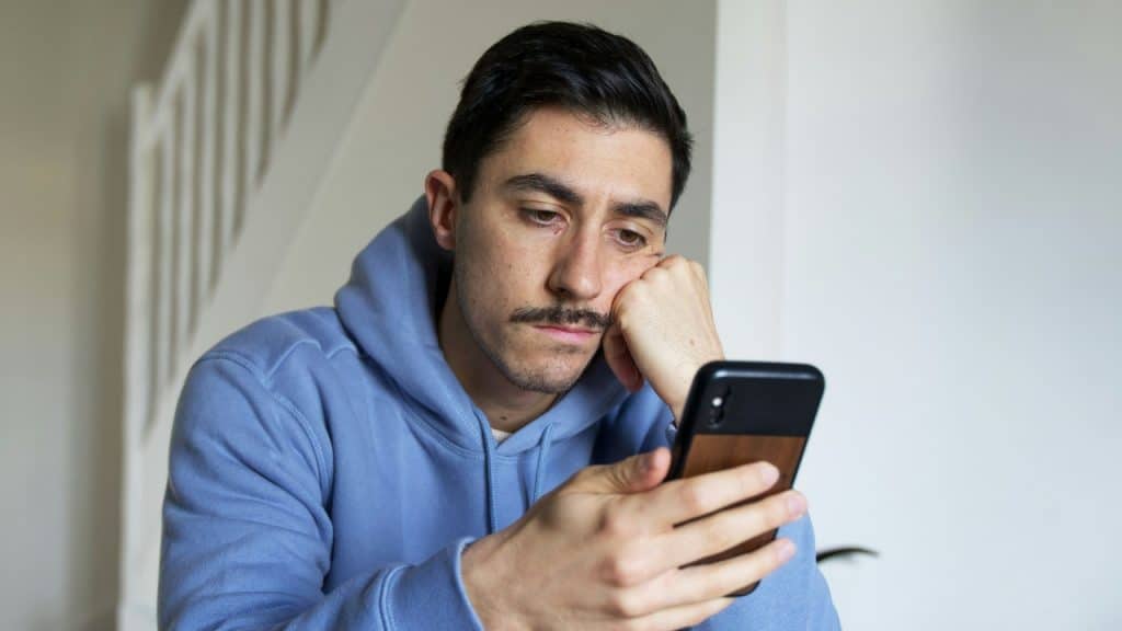 A man in a blue hoodie rests his head on his hand while looking at a phone.