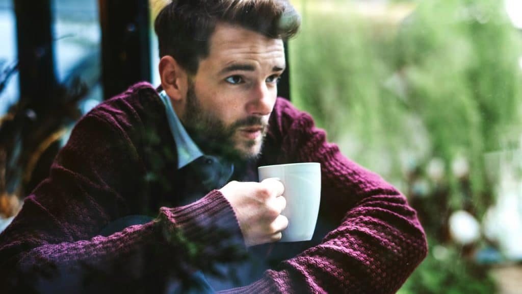 A man in a purple sweater looks out a window while holding a white mug.