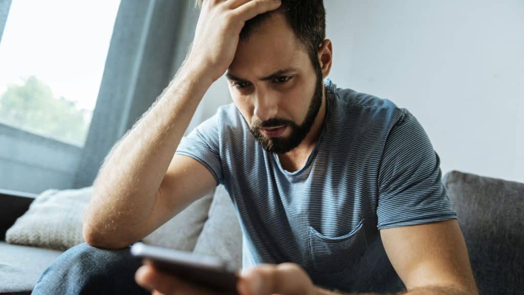 A bearded man in a striped shirt looking at a smartphone with a concerned expression.