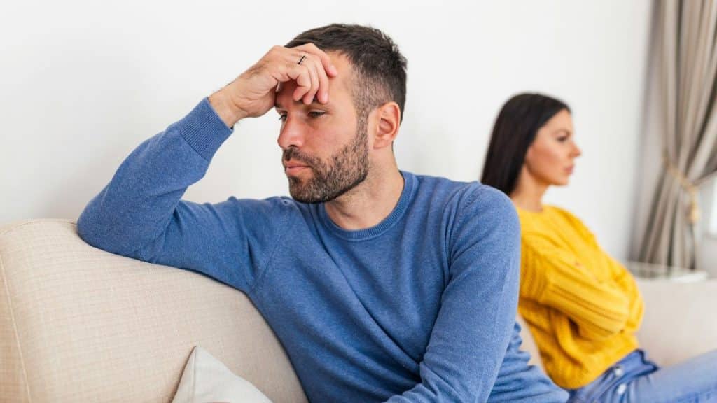 A man in a blue sweater touches his forehead while a woman sits behind him looking away.