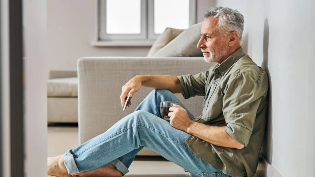 A grey-haired man in a green shirt and jeans sits on the floor near a sofa.