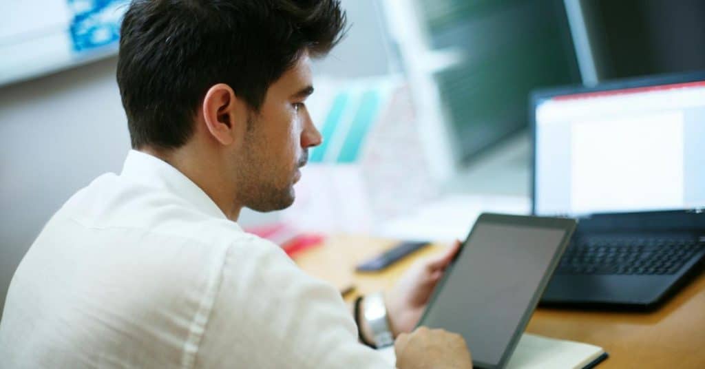 A man focused on a tablet while working at a desk with a laptop.