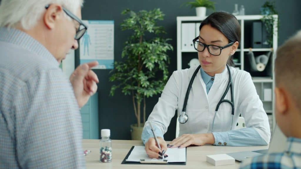 A female doctor in a white coat writes on a clipboard during a consultation.