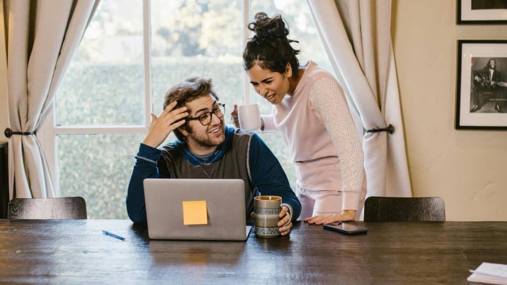 A woman standing beside a man working on his laptop as they talk and drink coffee.