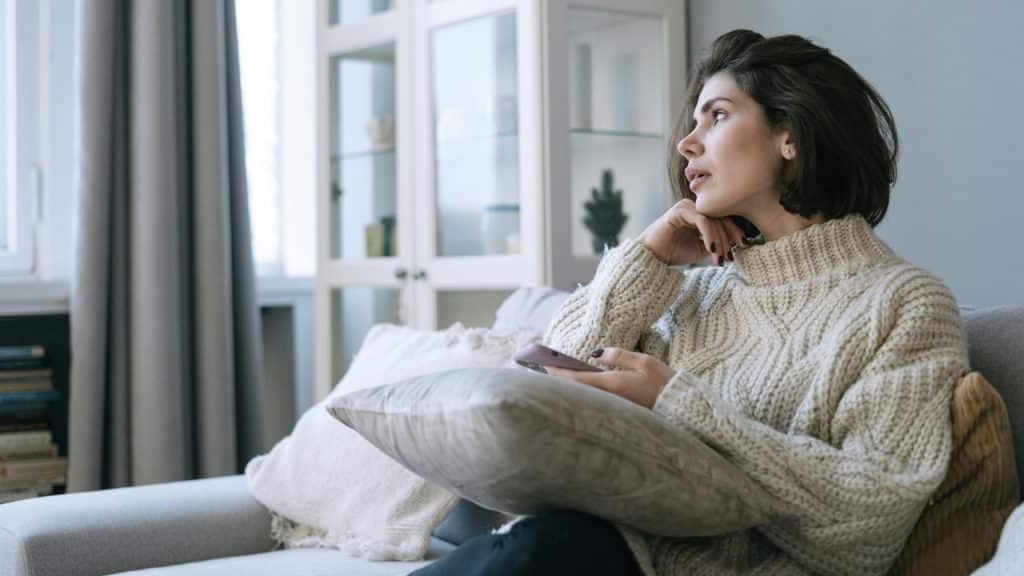 A woman in a white sweater sits on a grey couch holding a purple mobile phone.