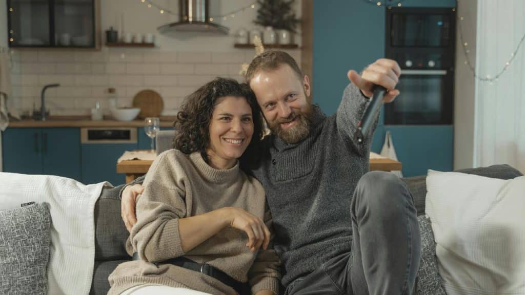 A smiling couple relaxing on a couch, the man holding a TV remote.