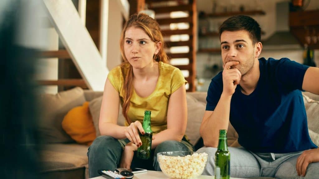 A man and woman sit on a couch holding bottles while watching a television screen.