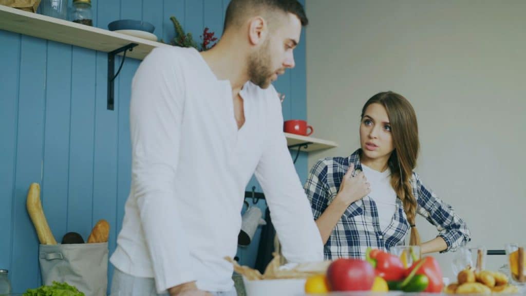 A man leans on a kitchen counter while a woman in a plaid shirt looks at him.