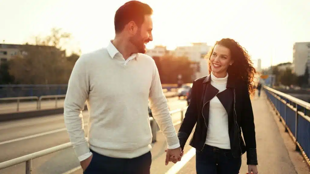 A man and woman hold hands while walking along a bridge during a bright sunset.