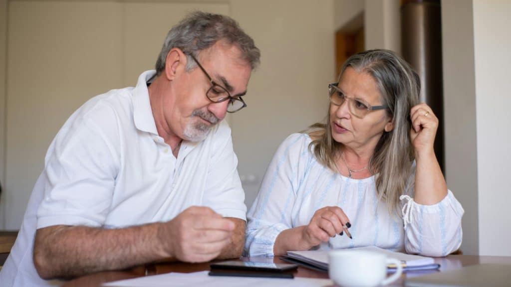 An older man and woman sit together at a table looking at papers and a tablet.