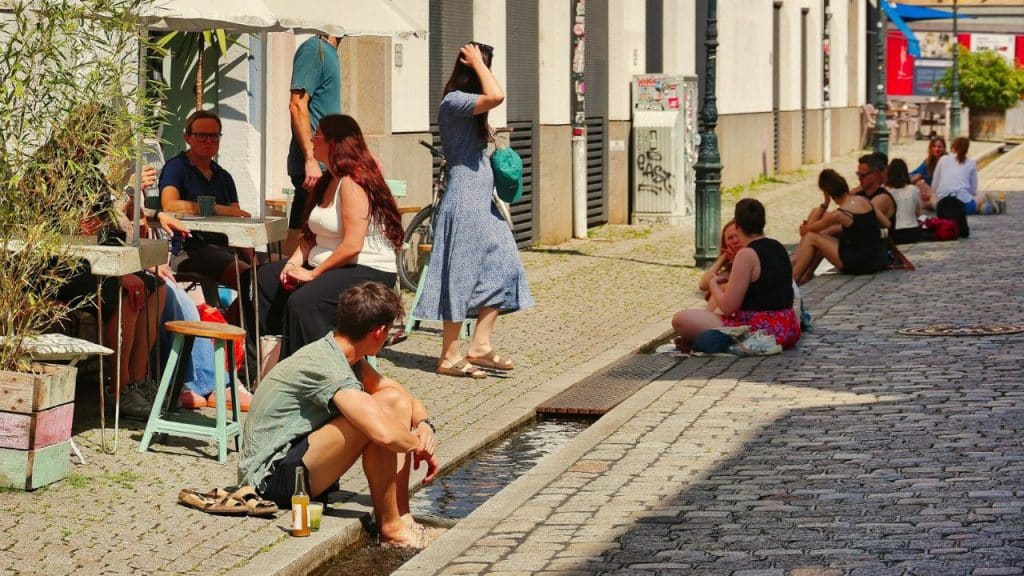 People sit at outdoor cafe tables and along a stone gutter on a sunny street.