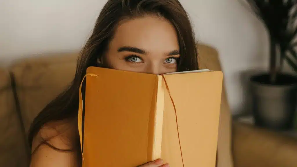 A woman holding a book up to her face while looking over it.