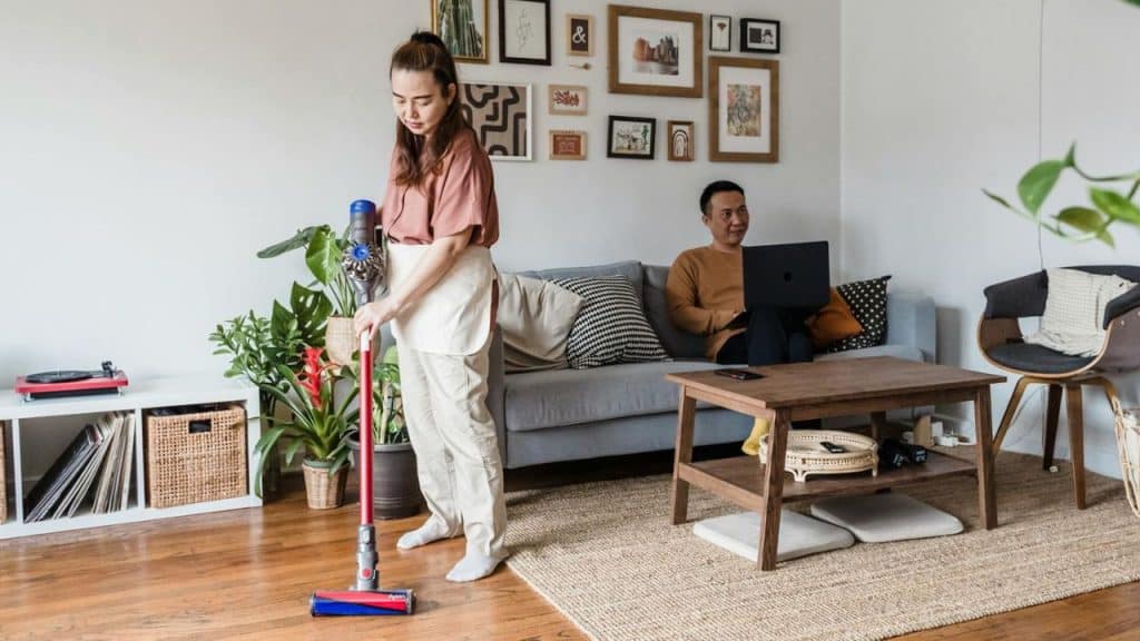 A woman vacuums a floor while a man sits on a sofa using a laptop.
