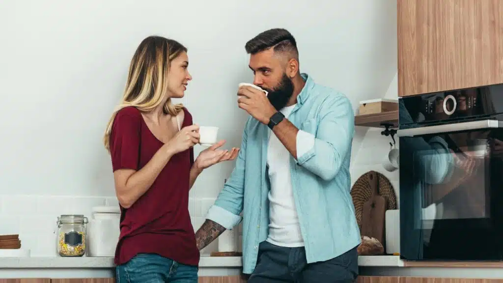 A man and woman stand in a kitchen, holding white mugs and talking to each other.