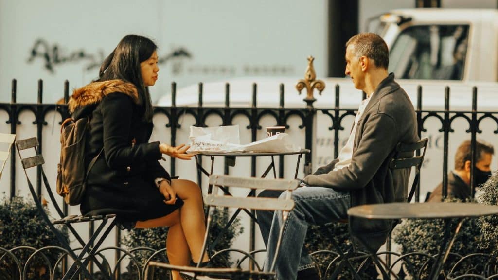 A man and woman sitting across from each other at an outdoor cafe table talking.