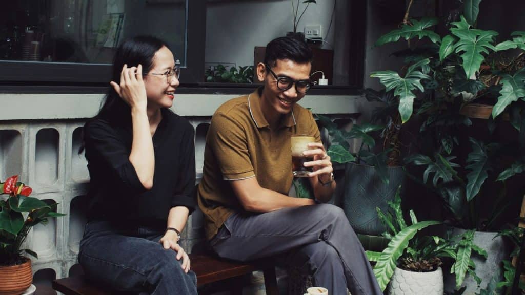 A man and woman sit on a bench outside, smiling and talking near potted plants.