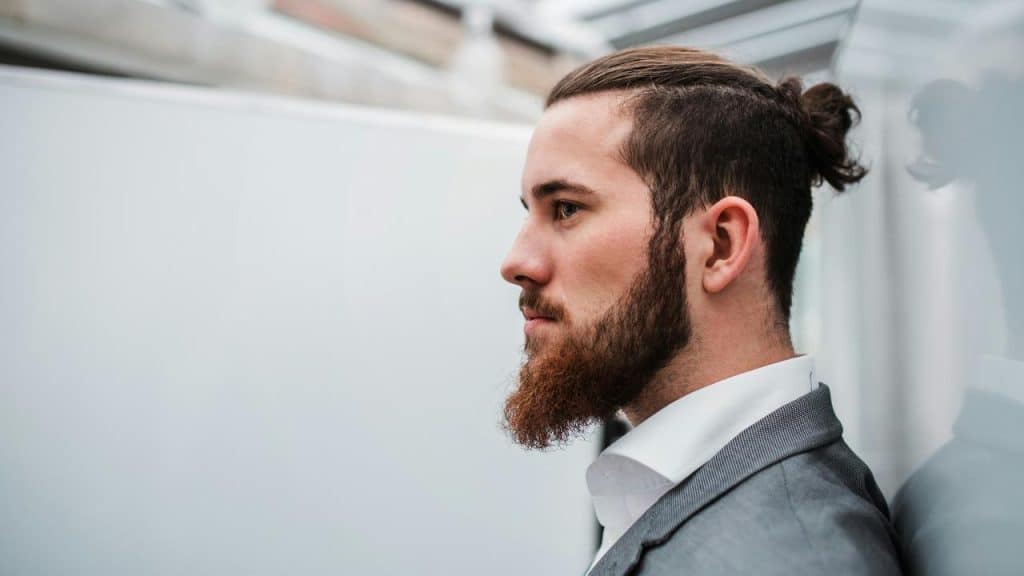 A man in a suit with a beard and tied-back hair standing sideways.