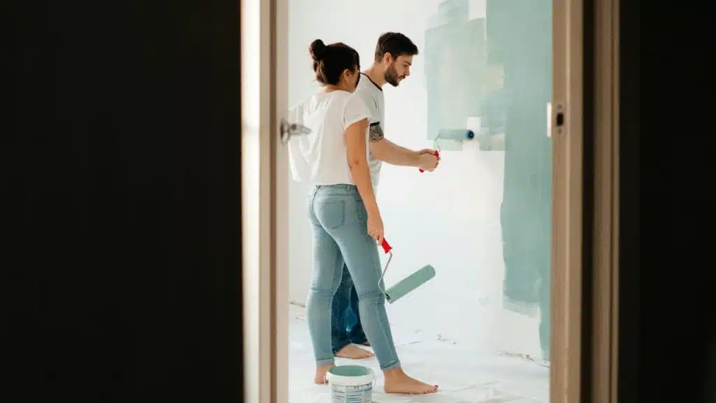 A man and woman stand together in a room, using rollers to paint a wall.