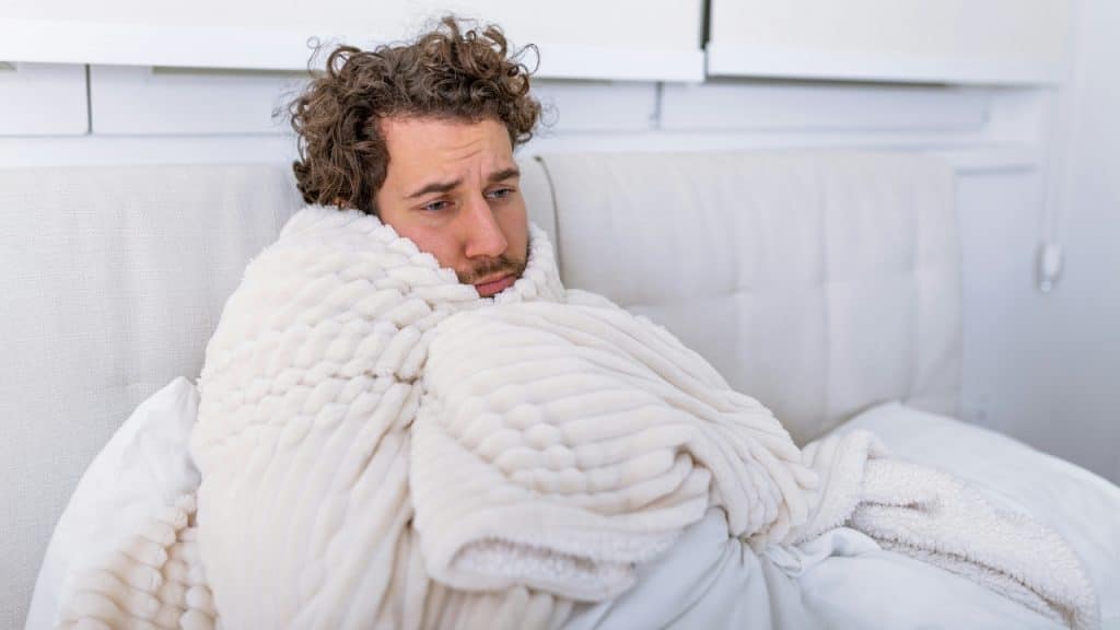 A man with curly hair sits in bed, wrapped tightly in a thick white blanket.