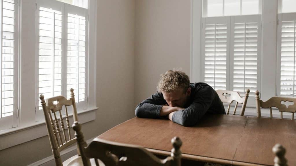 A man sits alone at a wooden table with his head resting on his arms.