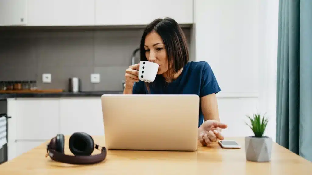 A woman drinks from a white mug while looking at a laptop on a wooden table.