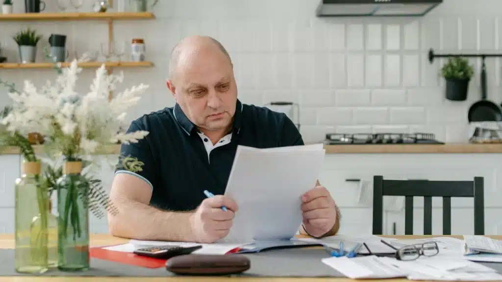 A man sits at a kitchen table holding a pen and carefully reading a paper document.