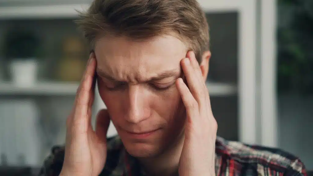 A man in a plaid shirt closes his eyes and rubs his temples with both hands.