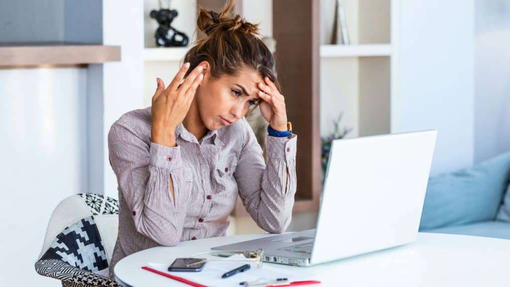 A woman sits at a white table with a laptop, resting her hands on her forehead.
