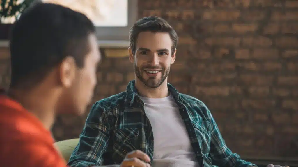 A smiling man in a plaid shirt talks to a person whose back is turned.
