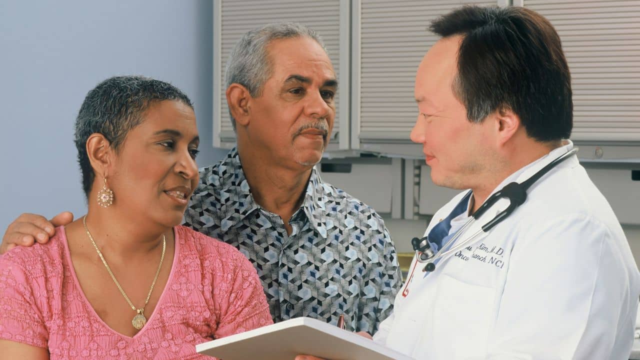 A man and woman in a medical office listen to a doctor holding a white clipboard.