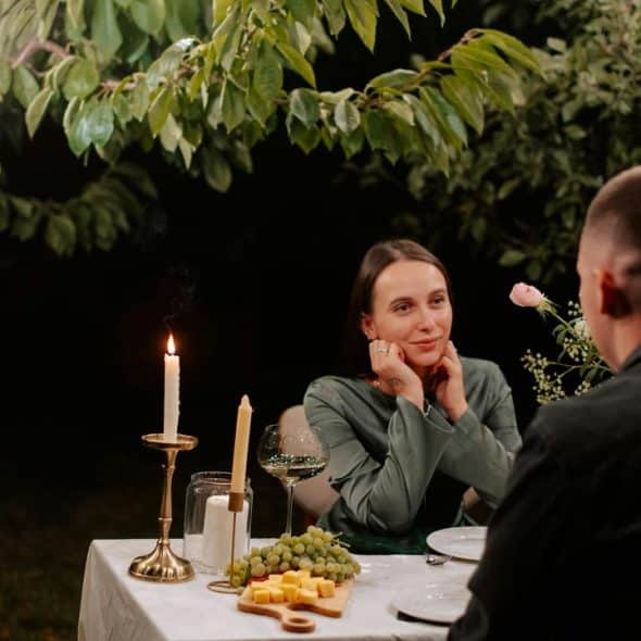 A couple enjoys a candlelit dinner outdoors under leafy trees at night.