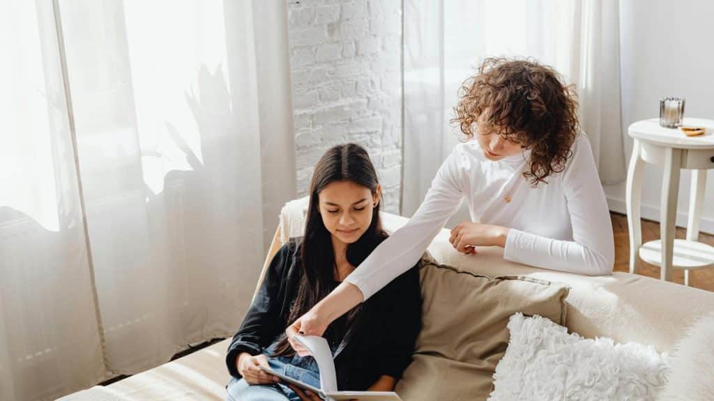 A woman helping her friend read a book on a cozy couch.